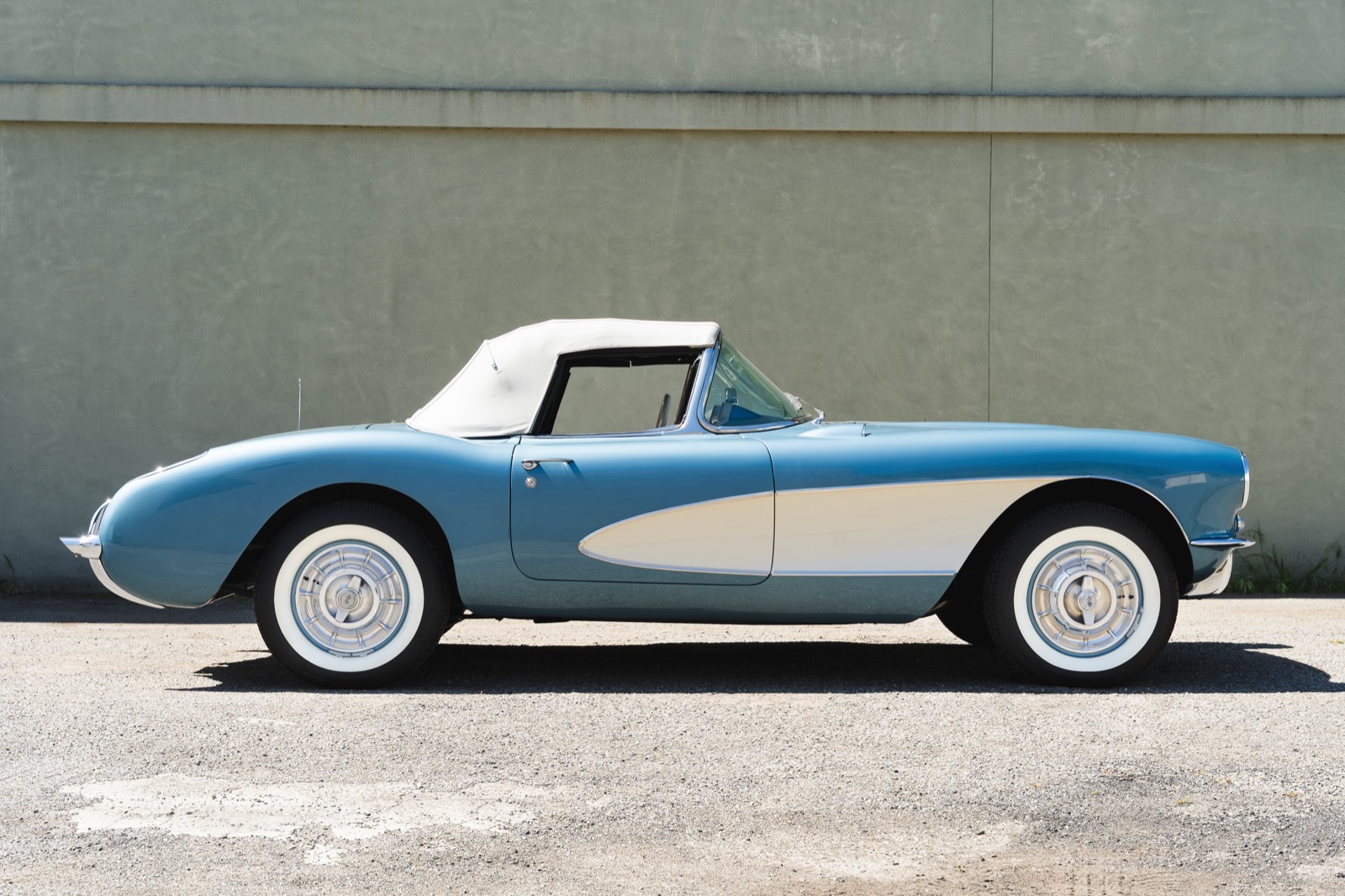 Side profile of a light blue 1957 Chevrolet Corvette with white side coves and whitewall tires, parked in front of a gray wall