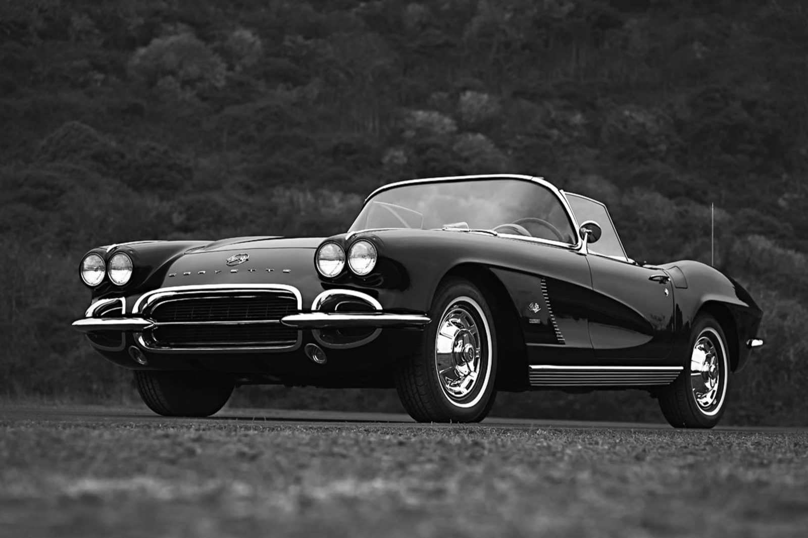 Black-and-white low angle of a black 1962 Chevrolet Corvette convertible with a brush-covered hillside behind it