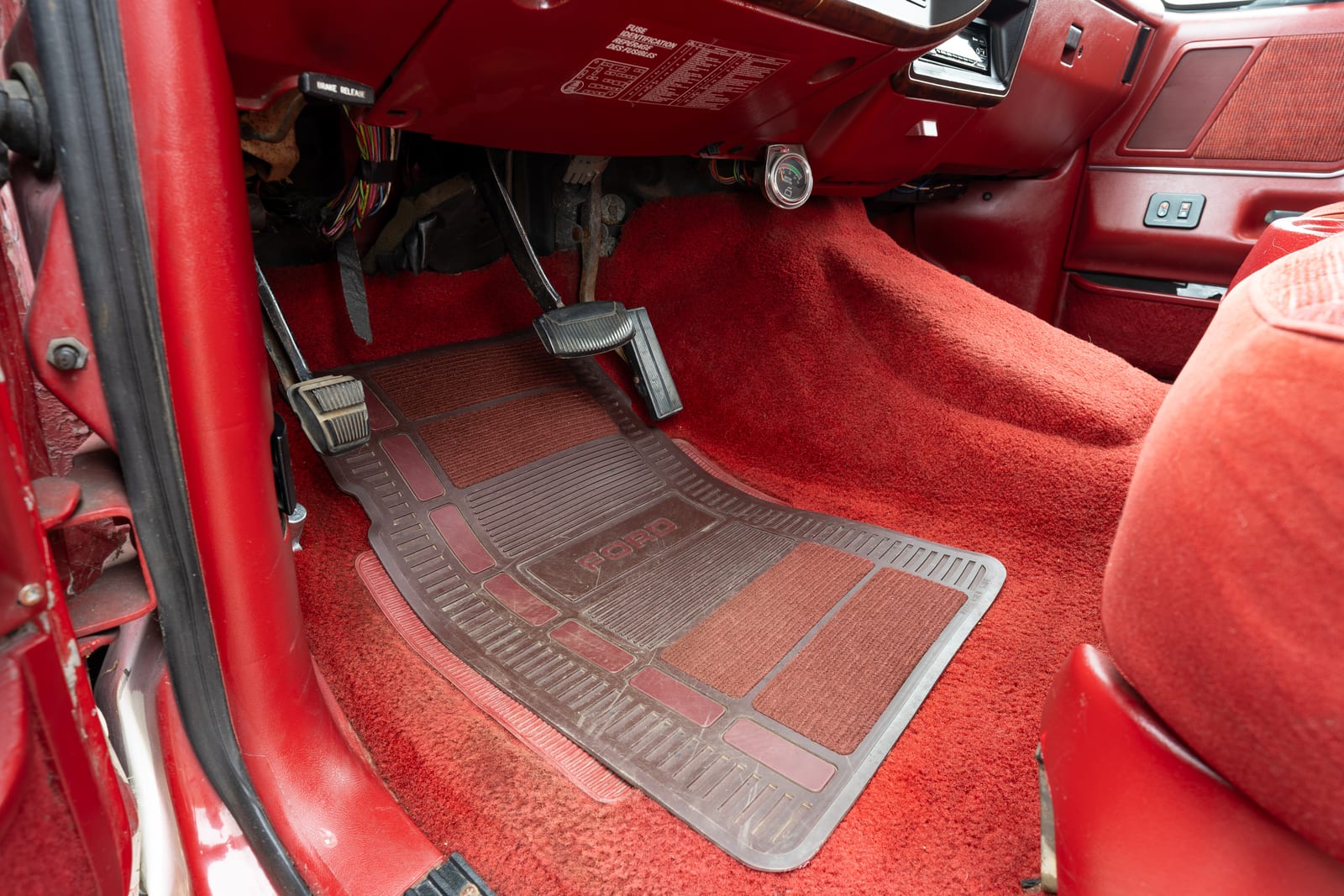Driver-side footwell of a 1989 Ford Bronco with a brown Ford rubber floor mat over red carpet, pedals and the underside of the dash above