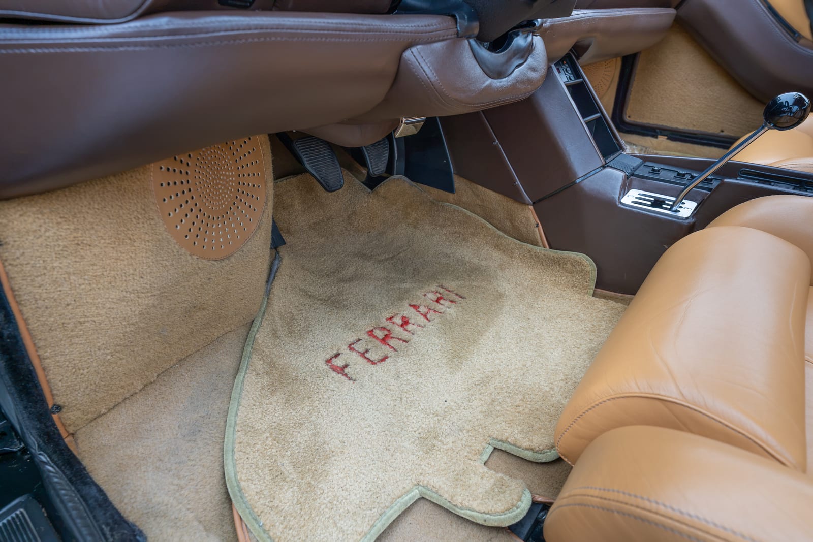 Tan carpet floor mat embroidered FERRARI in red on the driver side of a 1990 Testarossa, tan leather seat and pedals around it
