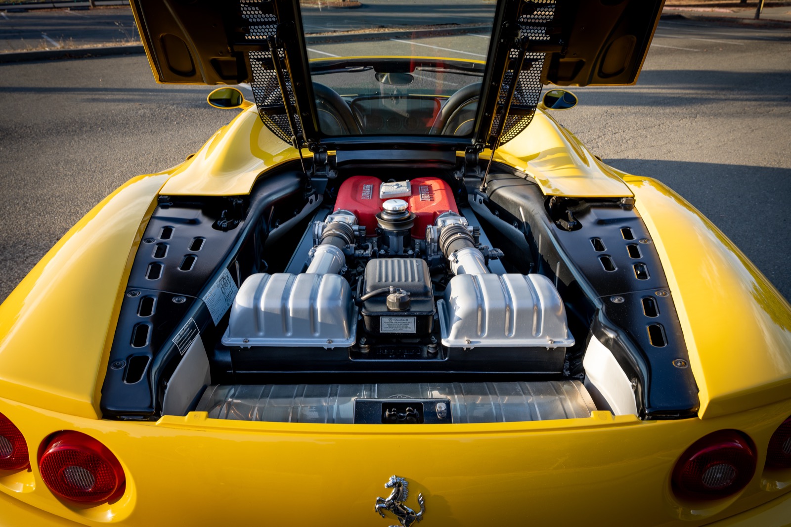 Rear view of a yellow 2004 Ferrari 360 Spider with the engine cover raised, showing the V8 engine and red intake manifolds