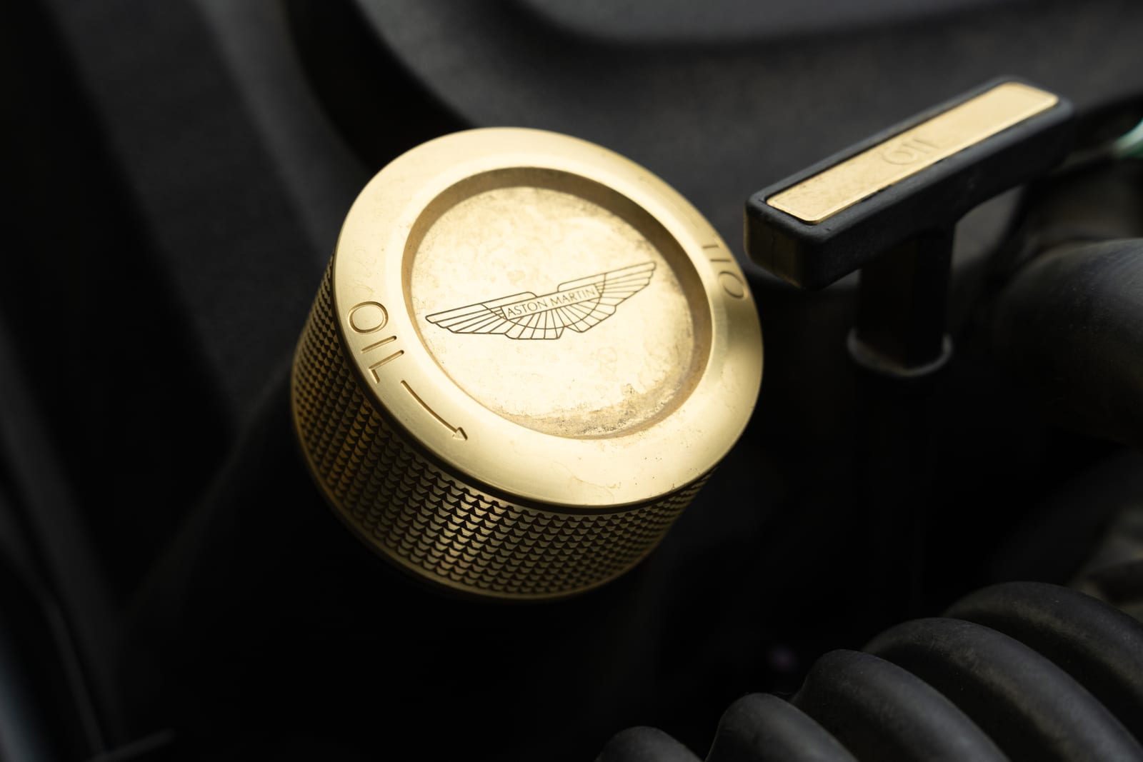 Gold anodised oil filler cap engraved with the Aston Martin wings in a 2017 DB11 engine bay, gold dipstick handle behind it