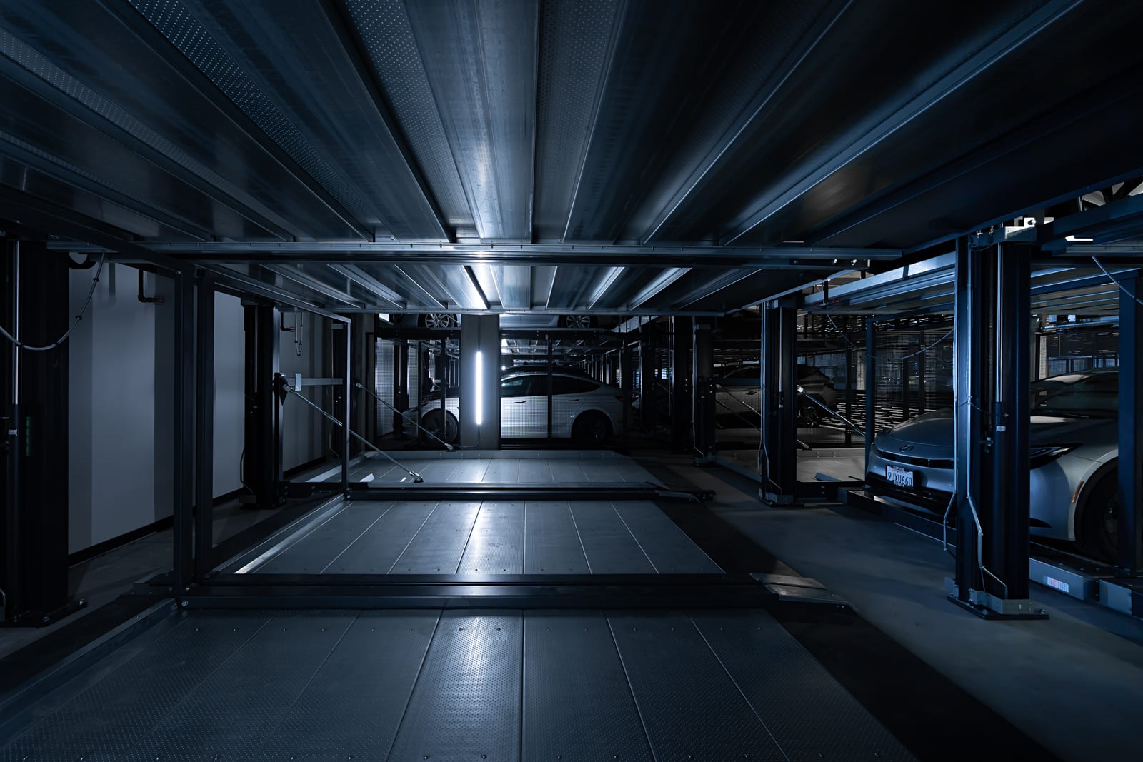 A dark blue-toned automated parking system lit by a single vertical light strip, cars on platforms receding into shadow