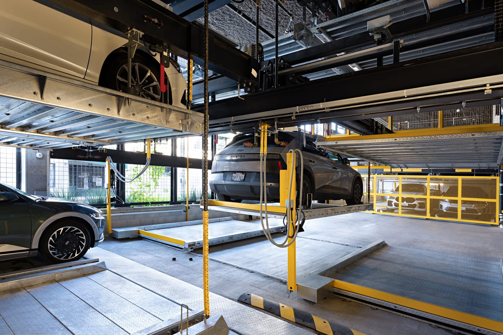 A car lifted on a steel pallet inside an automated parking system, daylight and greenery through the windows behind, a second car on the ground level