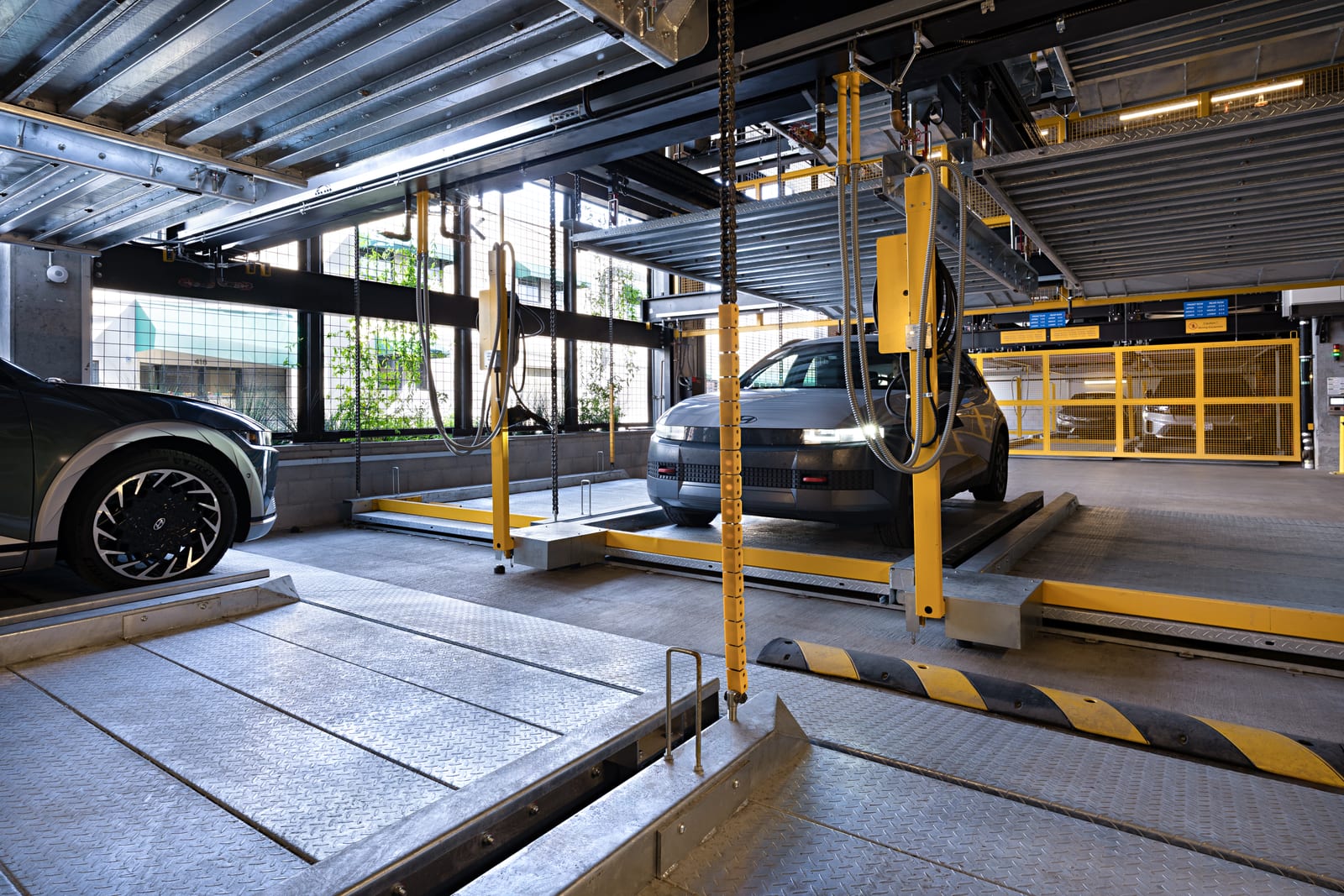 A car on a parking platform with headlights on, chains and yellow posts around it, bright daylight and trees through the windows behind