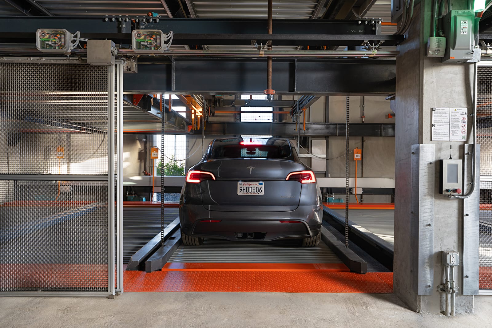 Rear view of a car with taillights lit, centered on a parking platform between steel posts and chains, orange floor plate in front