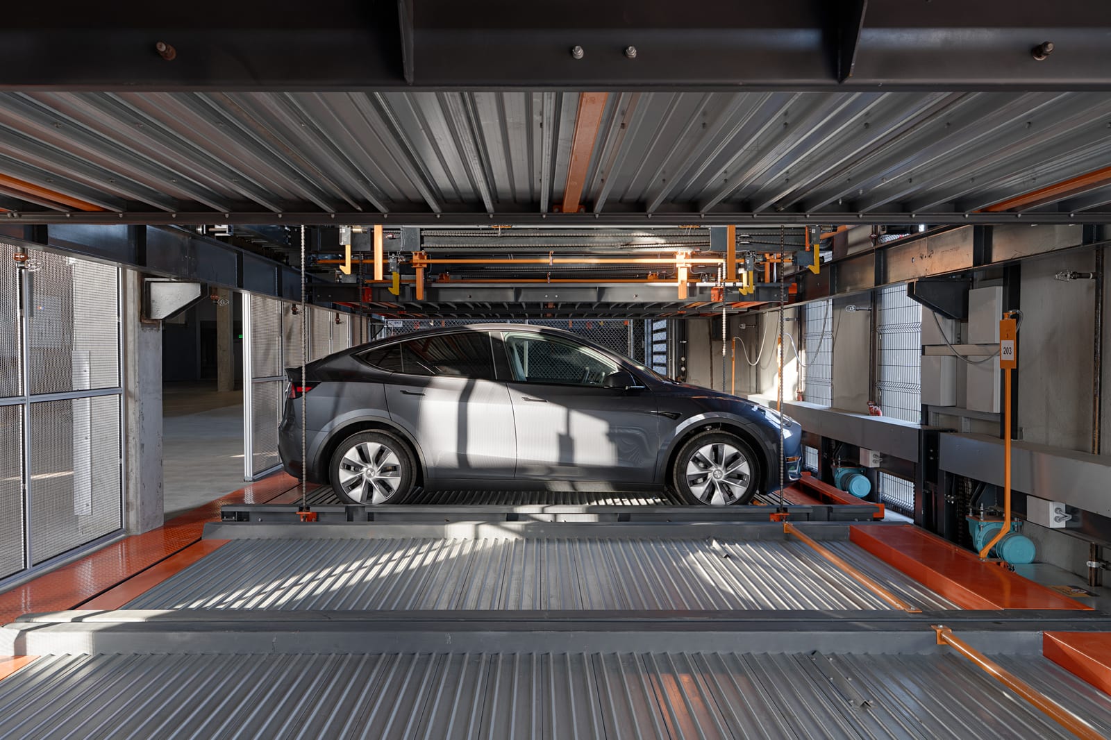 Low sun casting diagonal light and shadow across a car on a steel parking platform, orange rails and mesh walls around it