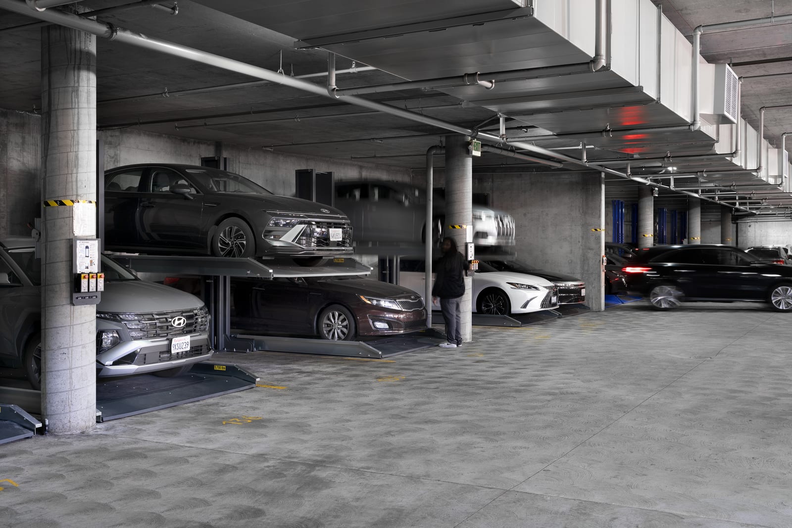 Cars blurred in motion past two-post parking lifts holding stacked cars, a technician standing at a concrete column