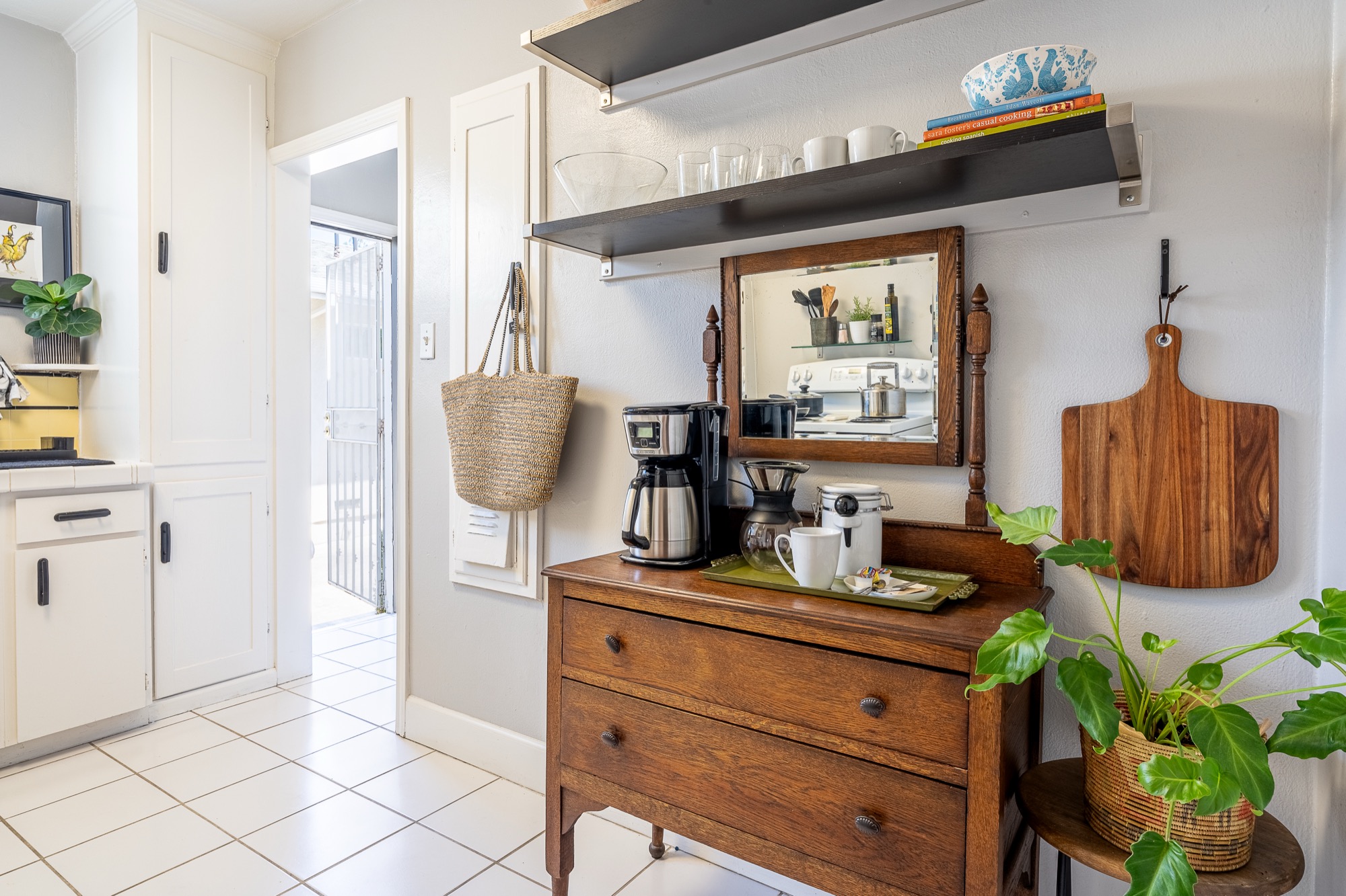 A vintage wooden dresser styled as a coffee station with a coffee maker, pour-over carafe, and mugs on open shelves, a mirror behind and a hanging cutting board beside it