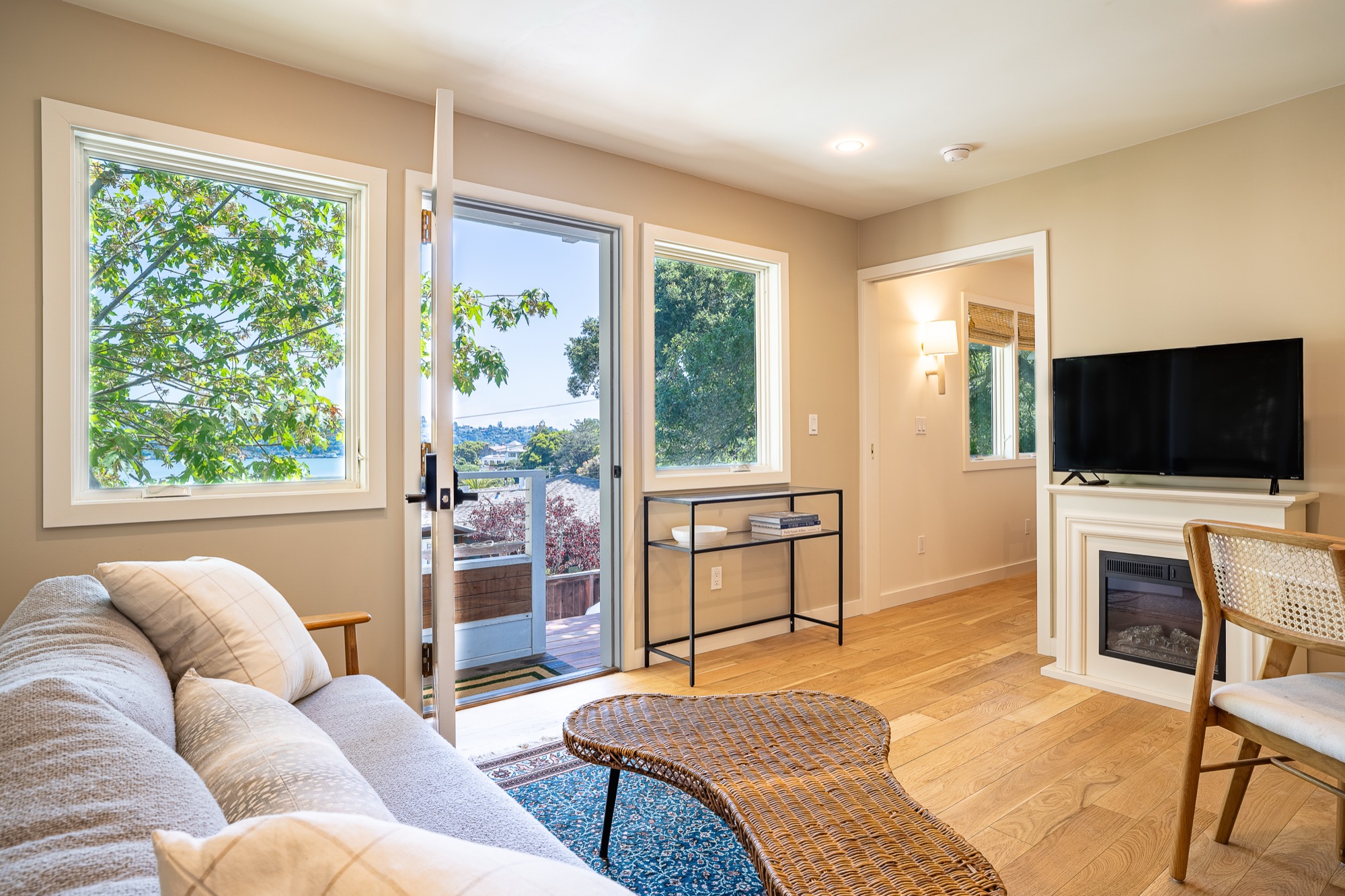 A living room with an open door and windows showing trees and water beyond, a light sofa in the foreground, wicker coffee table, and an electric fireplace under a TV