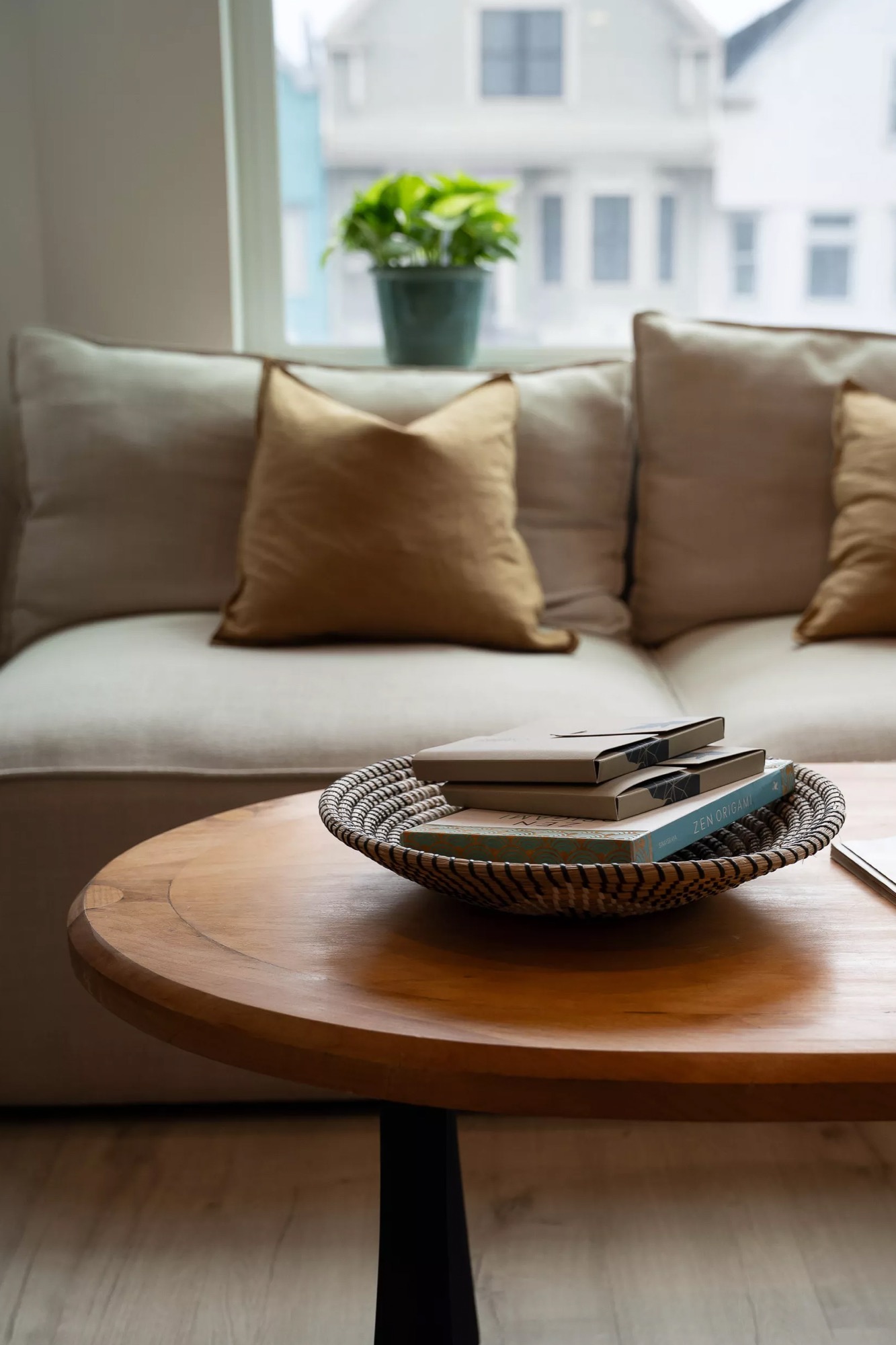 A close detail of a woven basket tray holding notecards and a Zen Origami book on a round wood coffee table, a linen sofa with gold pillows soft behind it