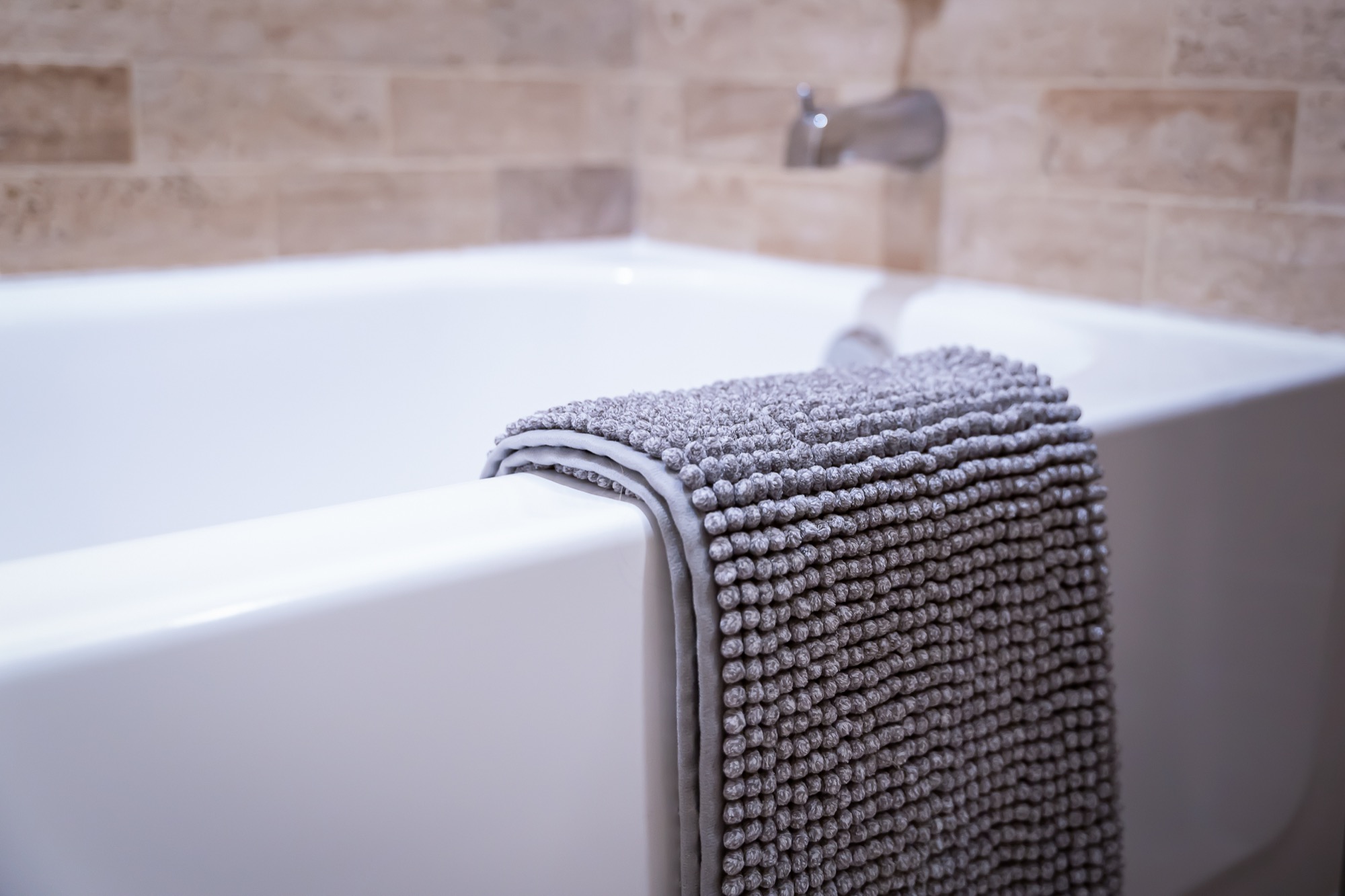 A close shot of a gray textured towel folded and draped over the edge of a white bathtub, travertine tile soft in the background