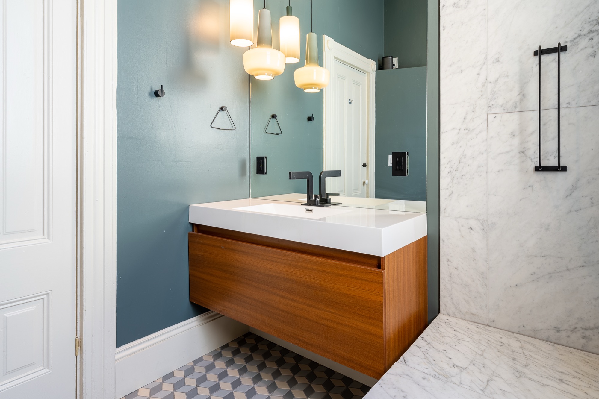A Victorian bathroom with teal walls, a floating wood vanity under pendant lights, black fixtures, and geometric cube-patterned tile flooring