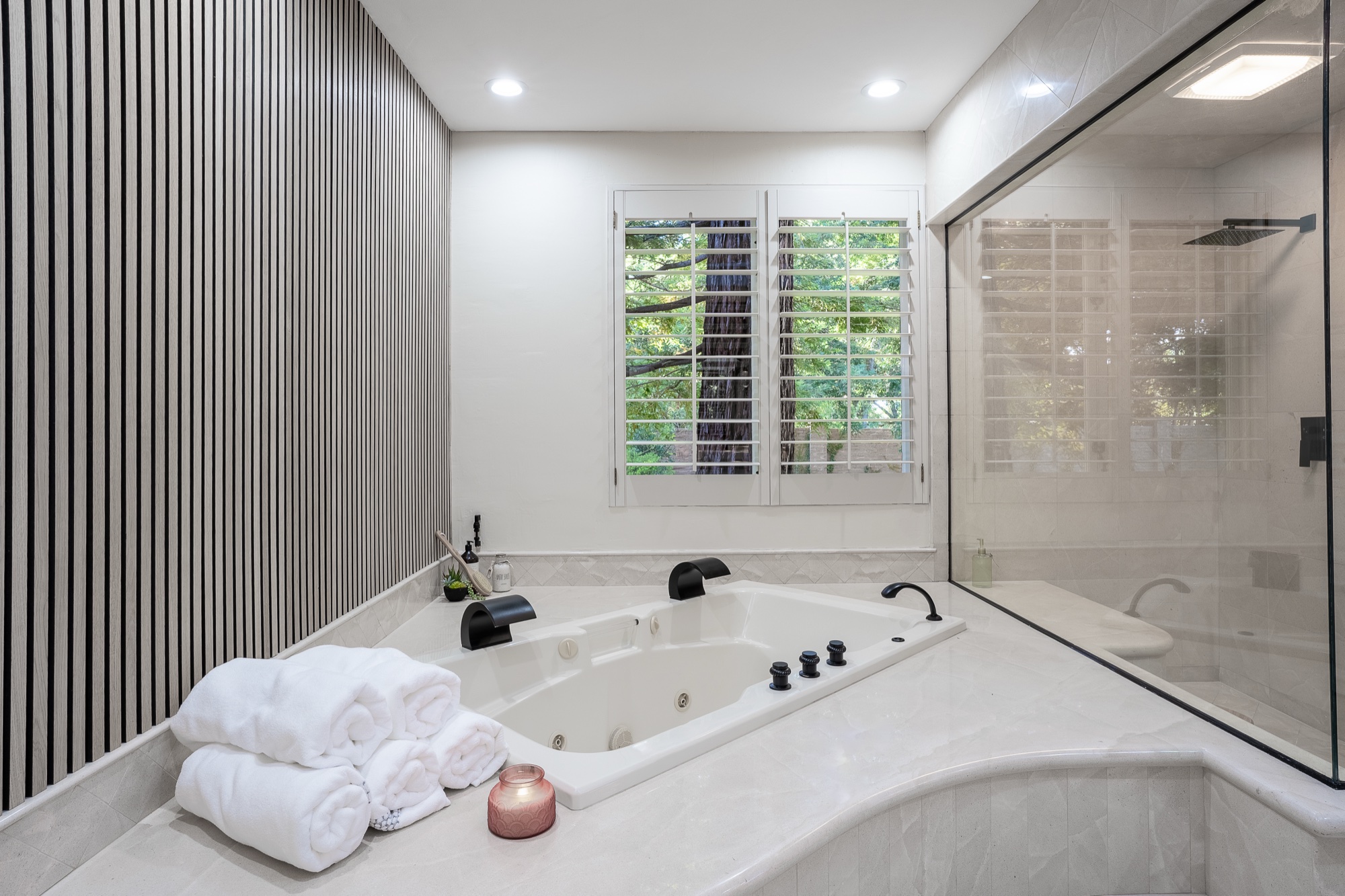 Rolled white towels stacked on the corner of a tub deck beside a jetted tub, with shuttered windows and a glass shower behind