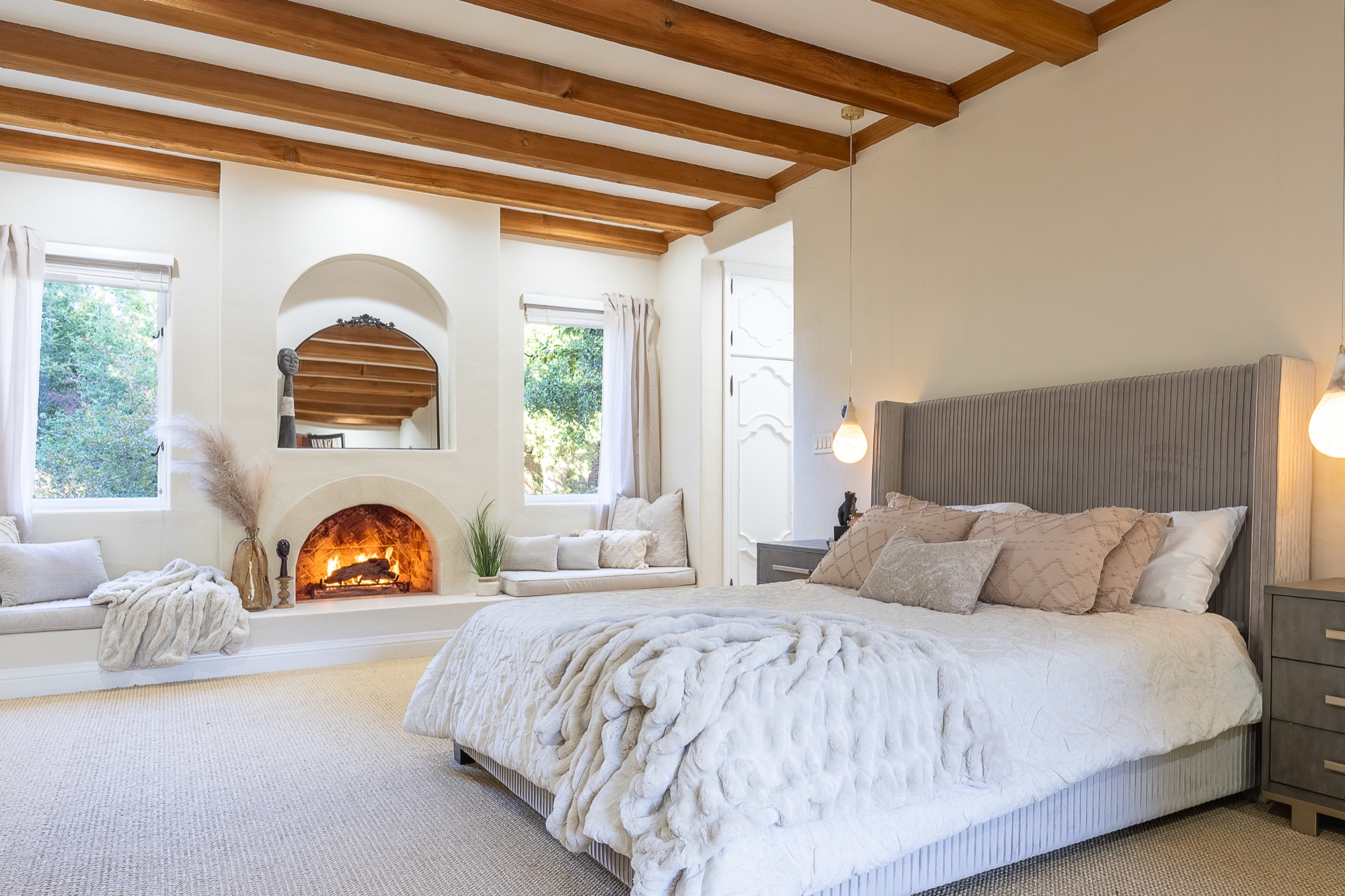 A styled bedroom with wood ceiling beams, a fur throw across the bed, window seats, and a lit arched fireplace