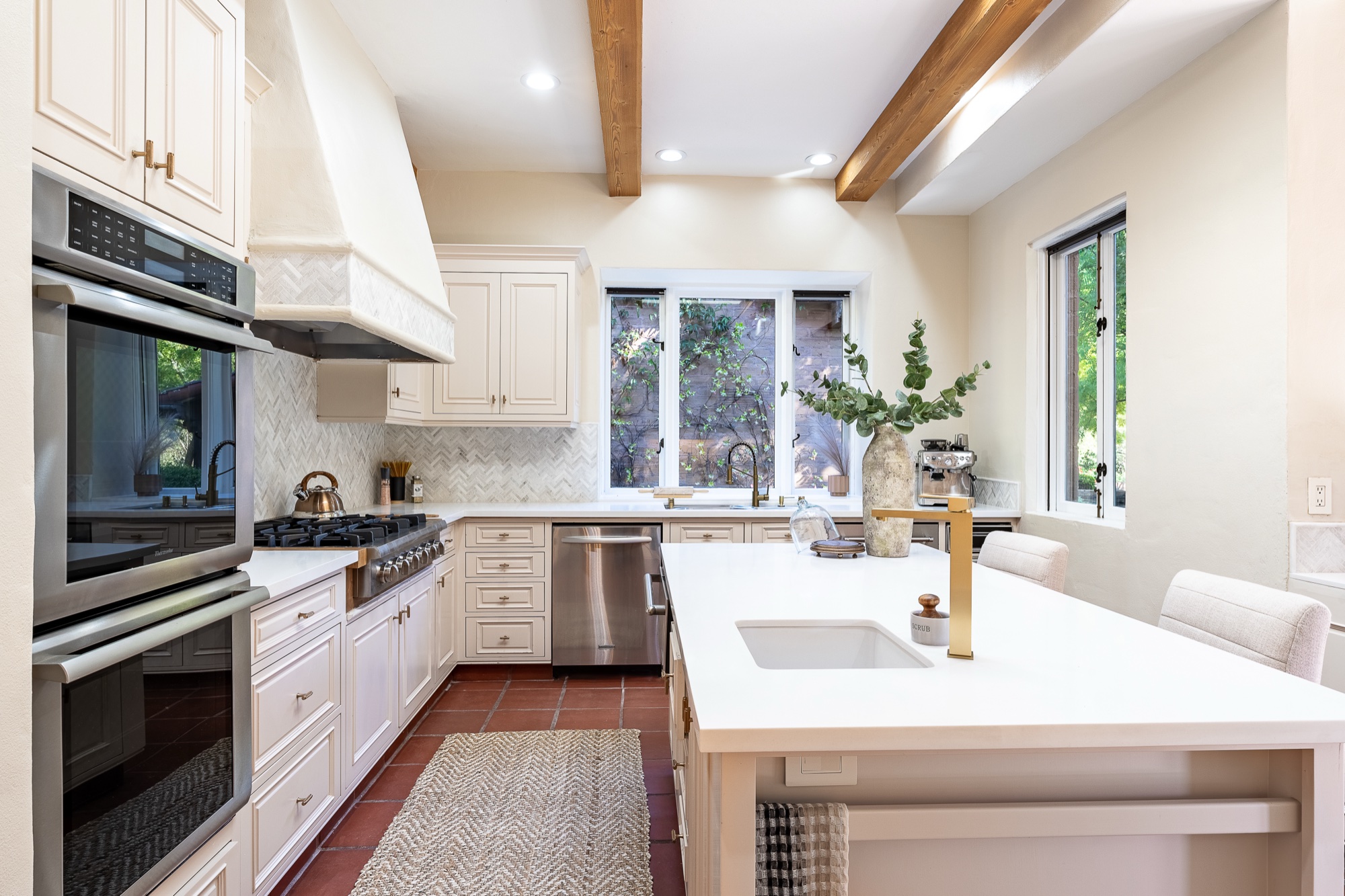 A white kitchen with wood ceiling beams, herringbone tile backsplash, a large island, and greenery through the windows