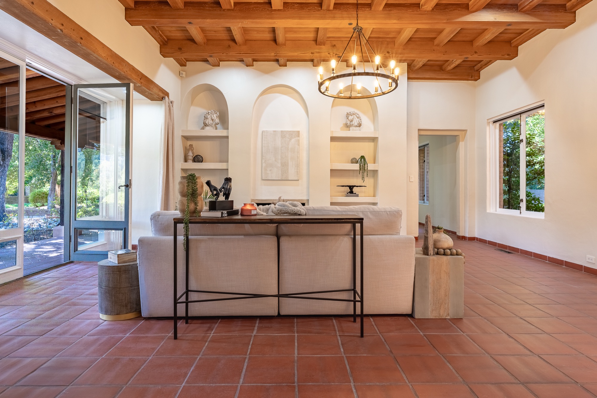 View from behind a cream couch and console table, facing arched wall niches and a fireplace, with a chandelier above and terracotta tile floors