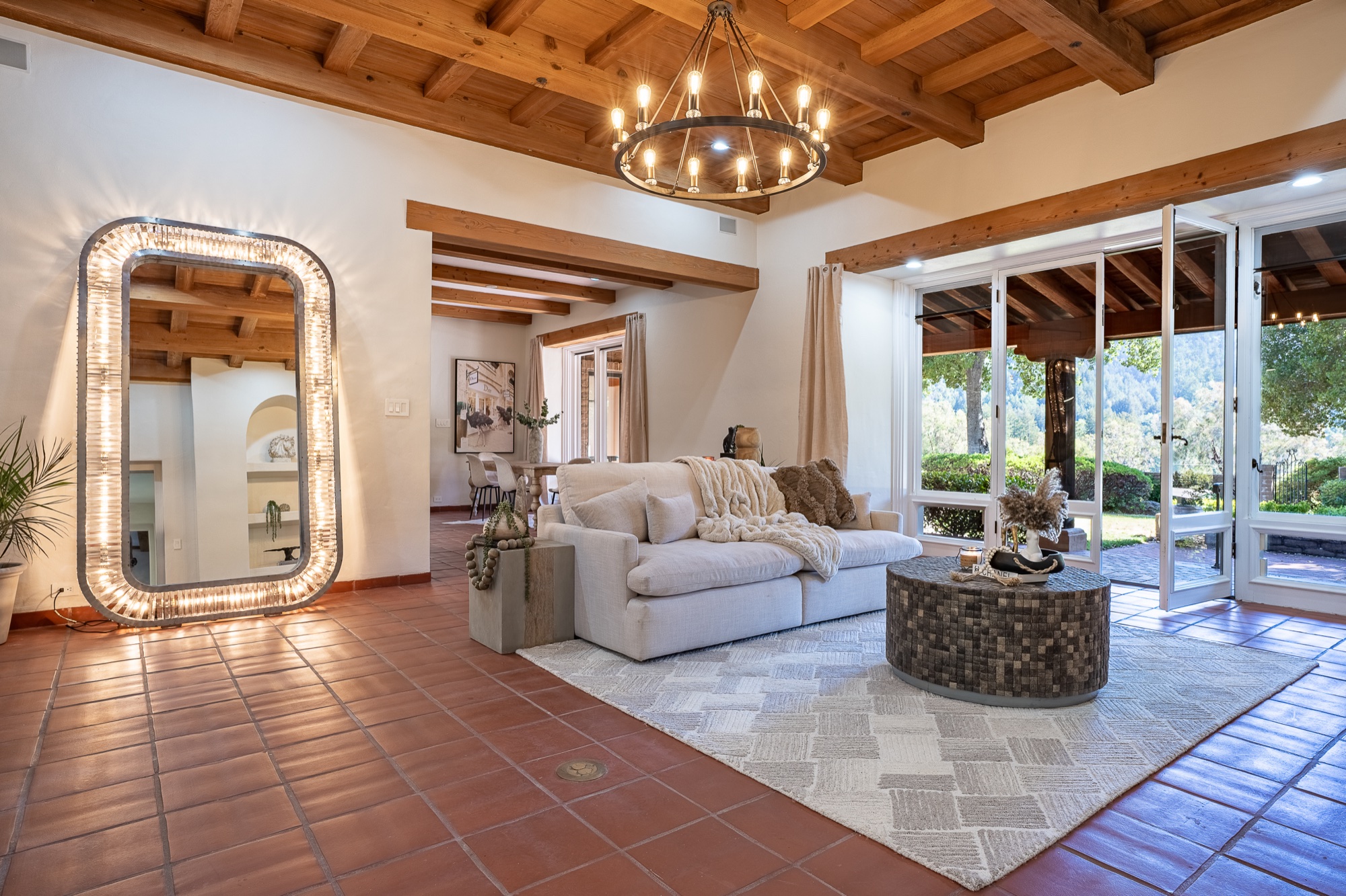 The same living room from the front: cream couch under a wagon-wheel chandelier, a lit mirror against the wall, and open glass doors revealing the backyard and wooded hills