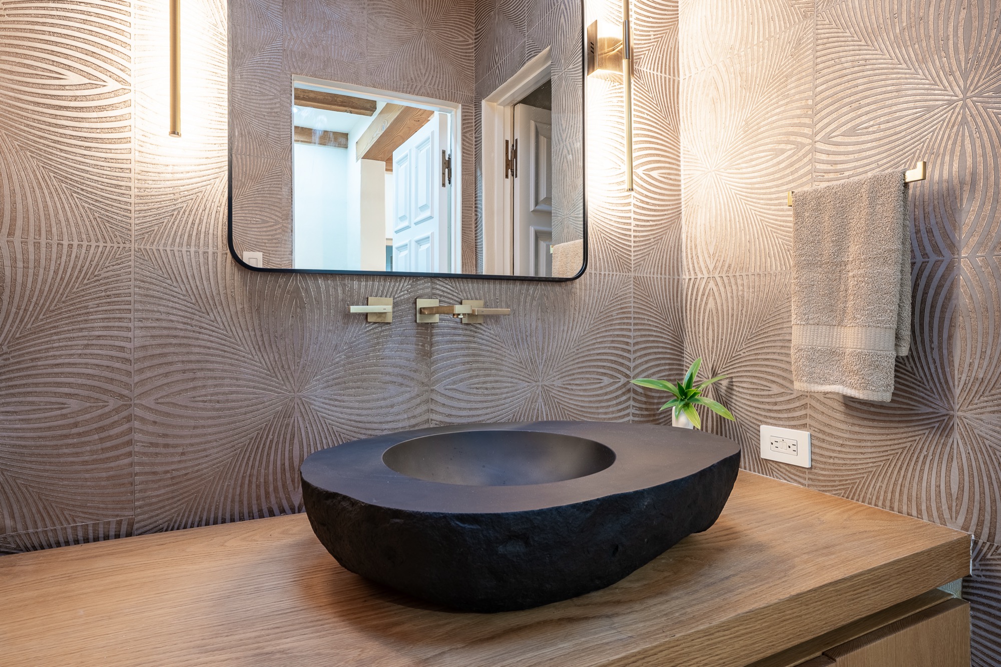 A carved black stone vessel sink on a light wood counter against patterned tile, brass wall-mounted faucet and vertical sconces beside a black-framed mirror